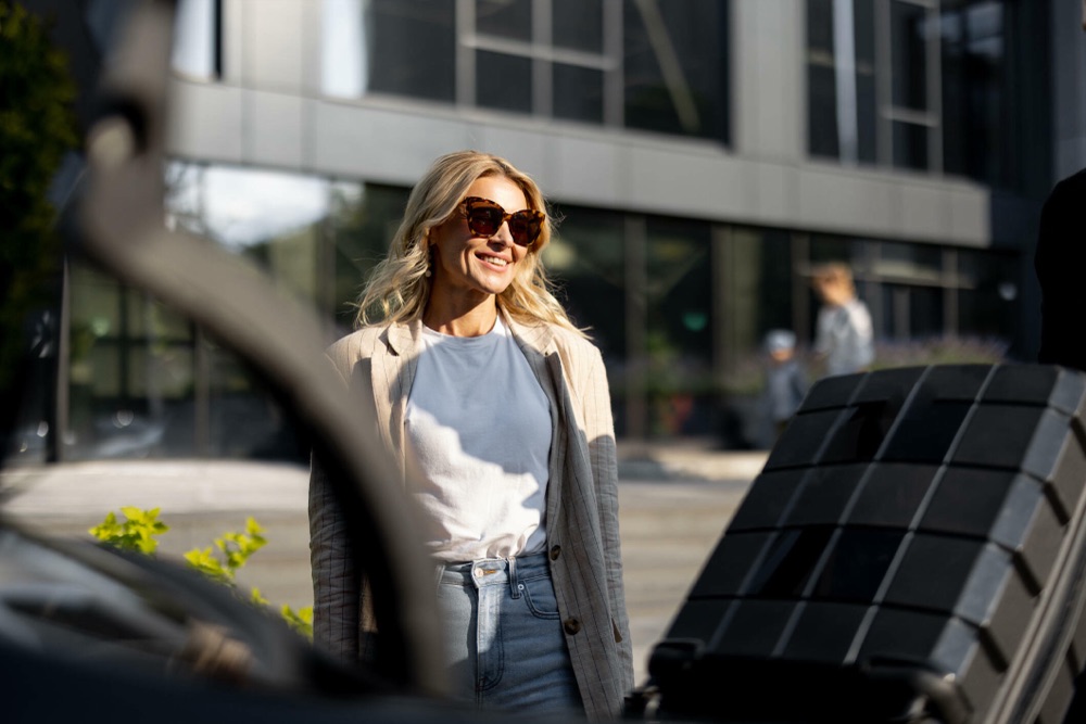 Traveller smiling beside a waiting car with luggage being loaded, as with a pre-booked Heathrow airport taxi instead of an Uber