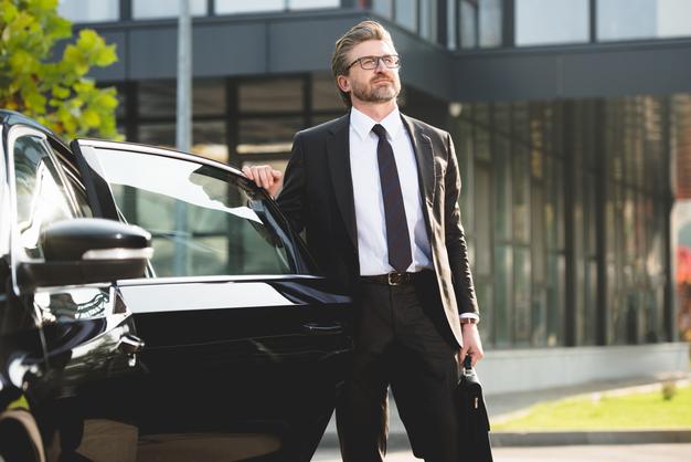 Professional taxi driver in a suit waiting beside an open car door, as a pre-booked driver waits for a passenger at Heathrow Airport